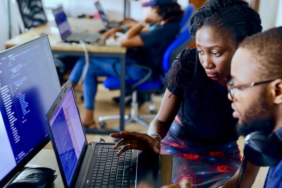 Deux collègues penchés ensemble sur un ordinateur portable affichant du code, dans un espace de travail partagé